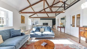 Ein großzügiges Wohnzimmer mit hoher Decke und freiliegenden Balken, einer blauen Sofagarnitur, einem Couchtisch und Blick in den Küchenbereich.