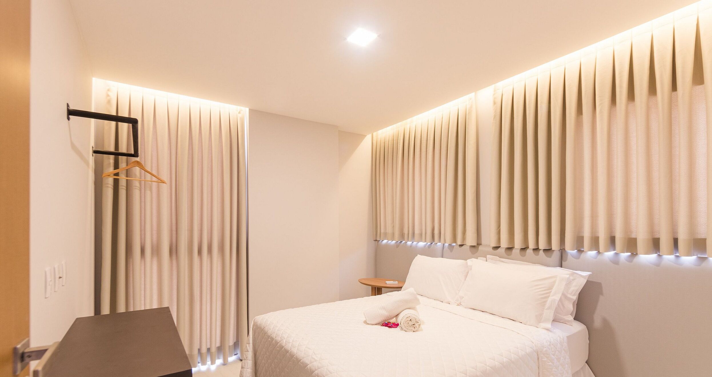 A modern hotel room with a neatly made bed, a hanging coat rack, and a small bedside table.