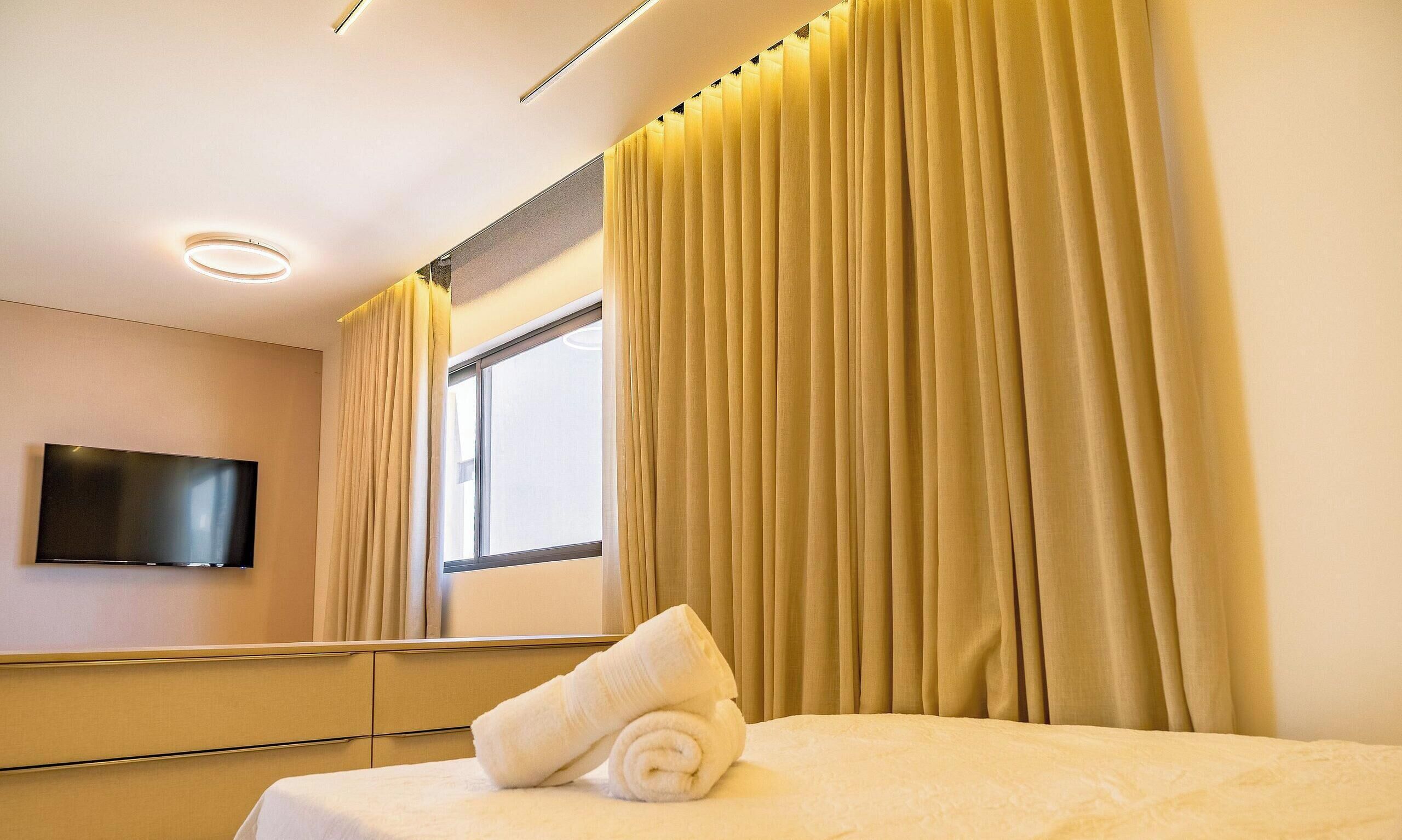 A hotel room with a bed, a flat-screen TV mounted on the wall, and a window with curtains.