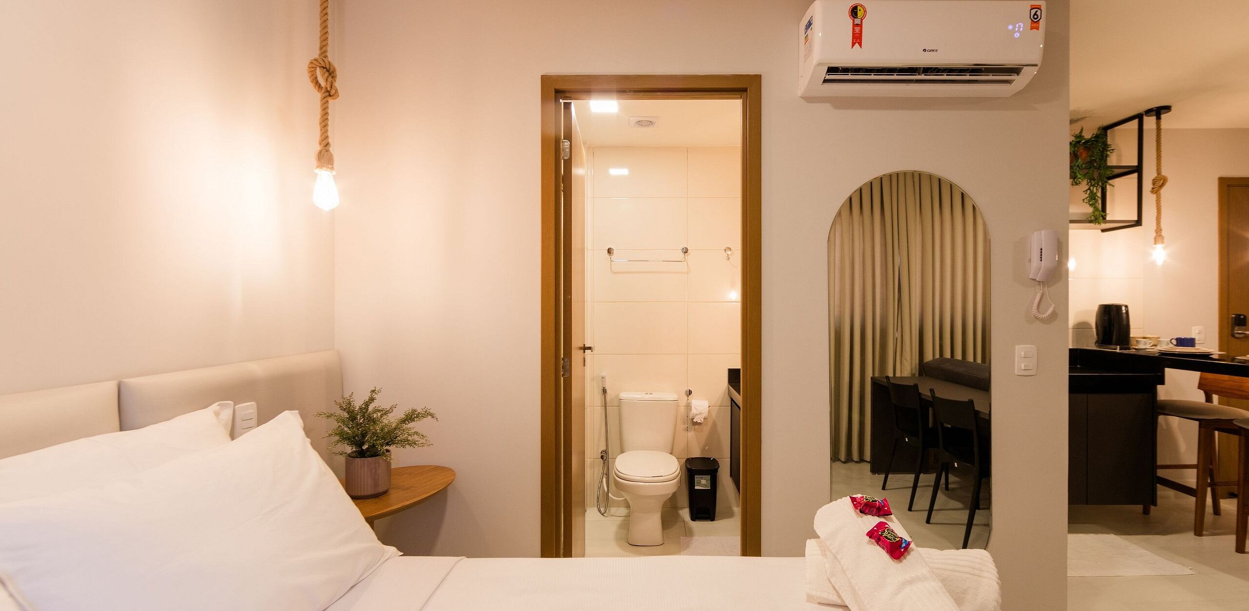 A hotel room with a bed, a small bedside table with a plant, a bathroom with a toilet and shower, and a kitchenette area.