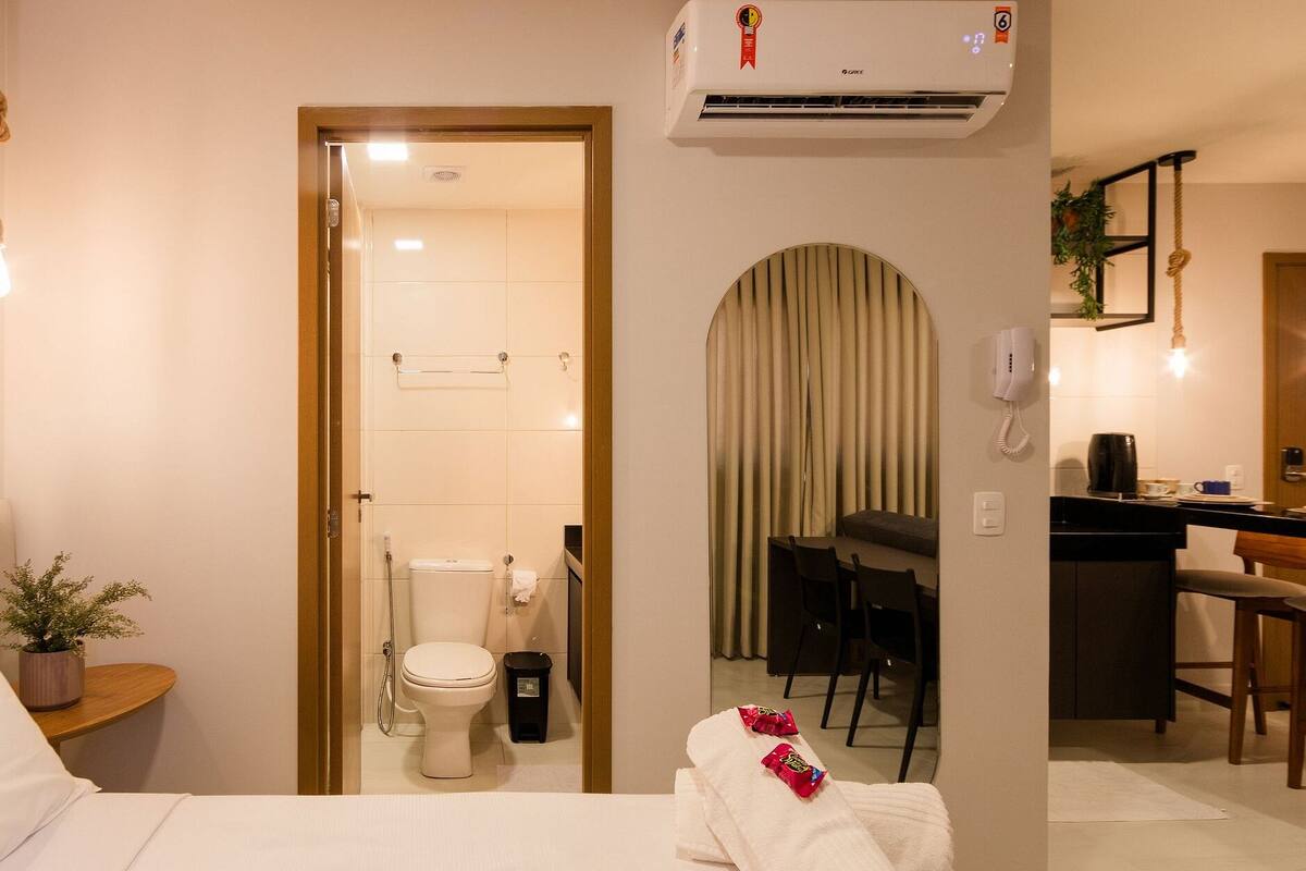 Une chambre d’hôtel comprenant un lit, une petite table de chevet avec une plante, une salle de bain avec des toilettes et une douche, ainsi qu’un coin cuisine.