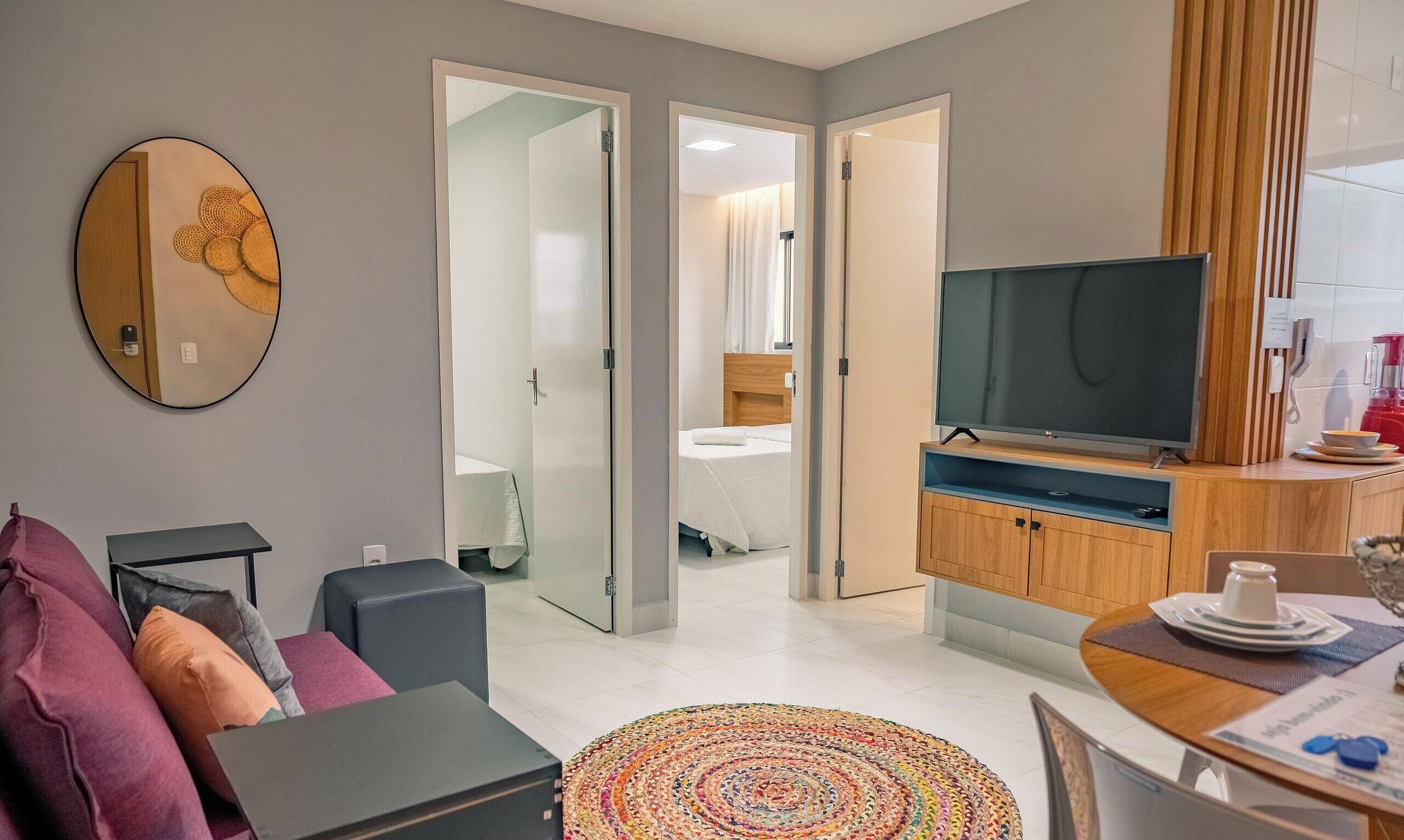 A modern hotel room with a sofa, a TV, a dining area, and a bathroom visible through an open door.