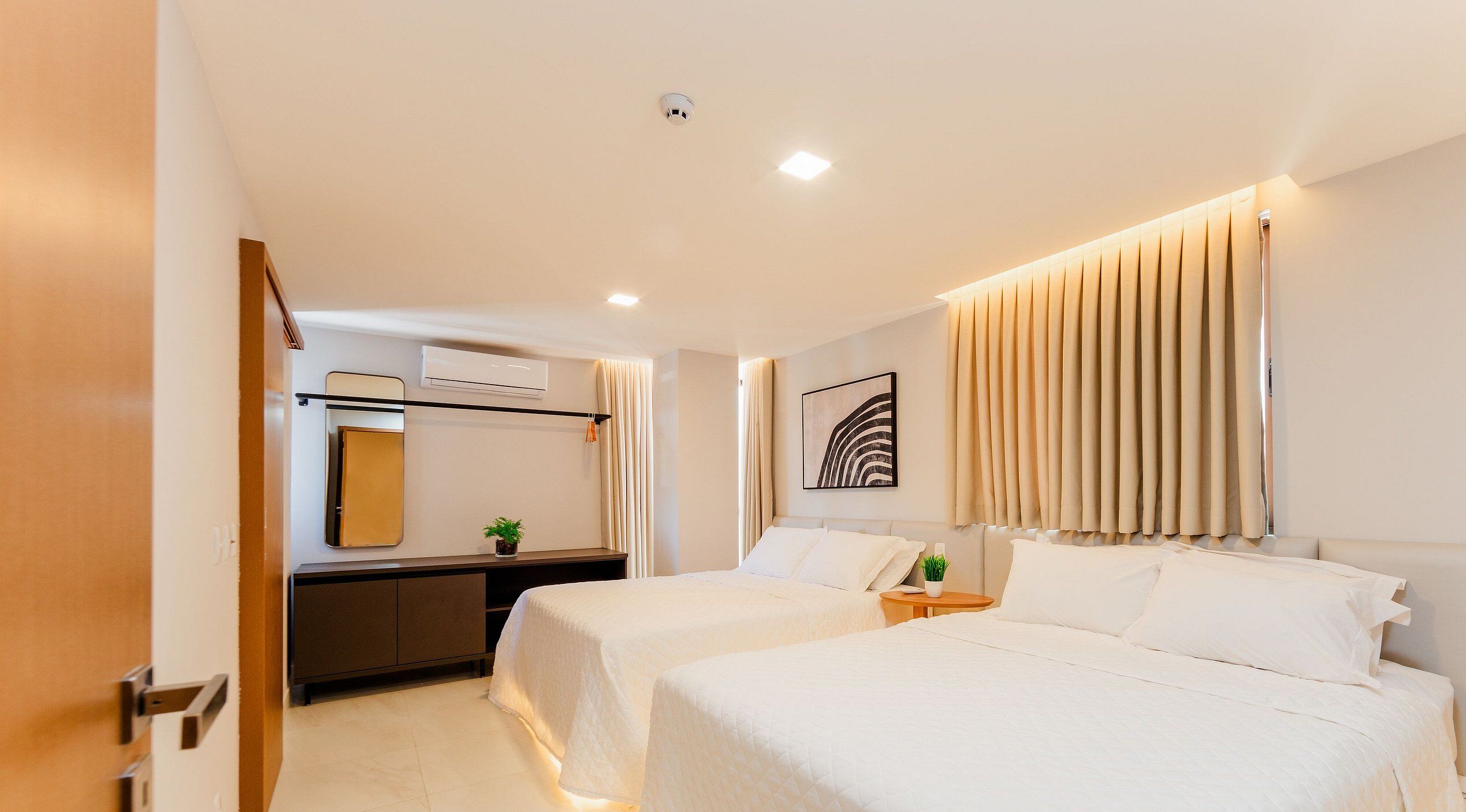 A hotel room with two beds, a desk, a mirror, an air conditioning unit, and a window with curtains.