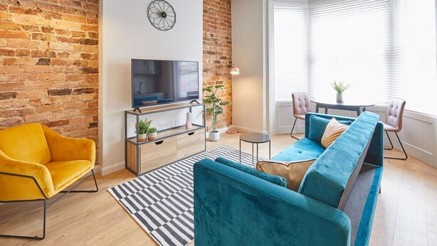 A modern living room with a brick wall, a flat-screen TV, a sofa, a chair, and a dining area with a table and chairs. - Host Stay Anchor Apartment North Marine House (Scarborough)