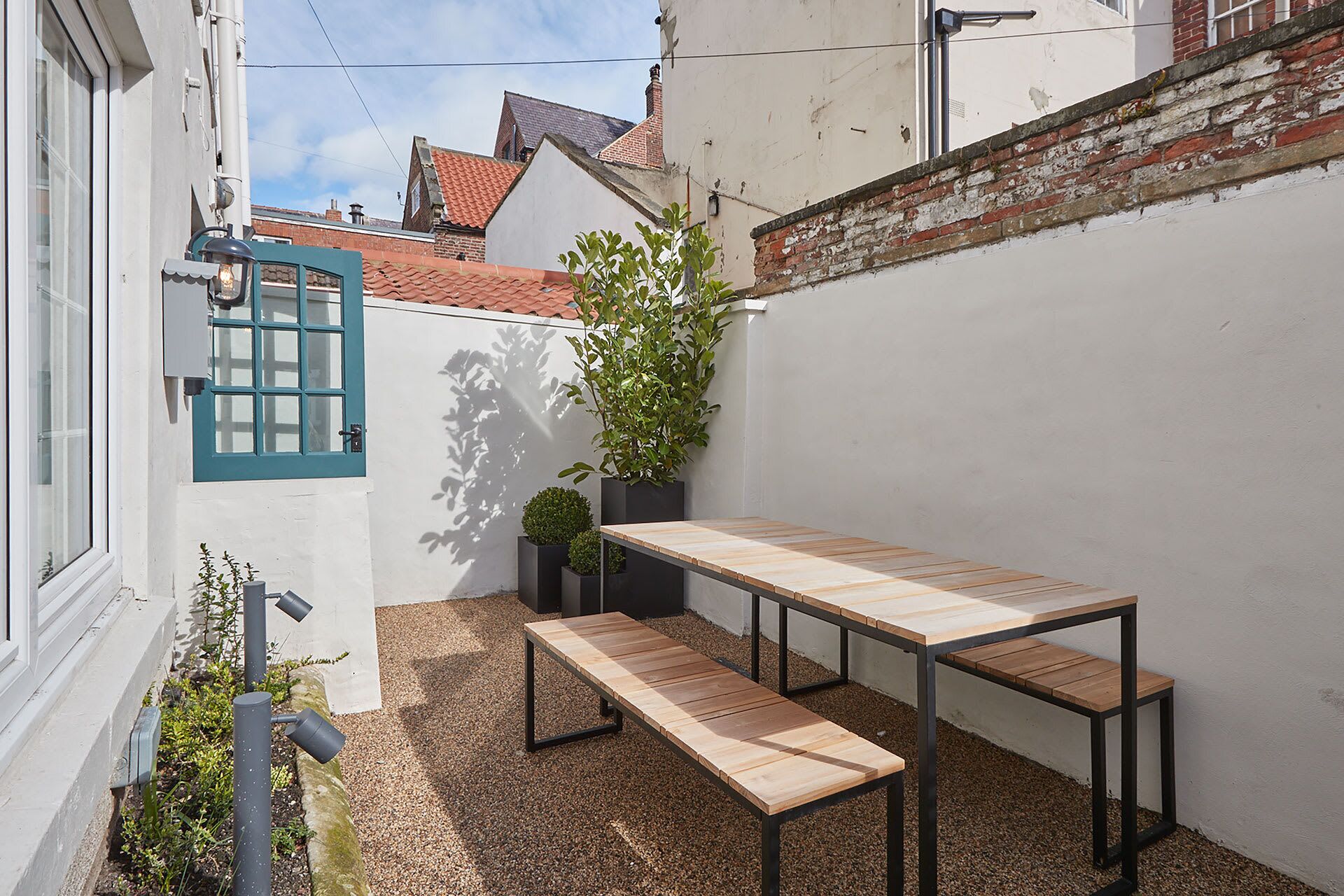 A small outdoor seating area with a wooden table and benches, surrounded by a white wall and a brick wall.