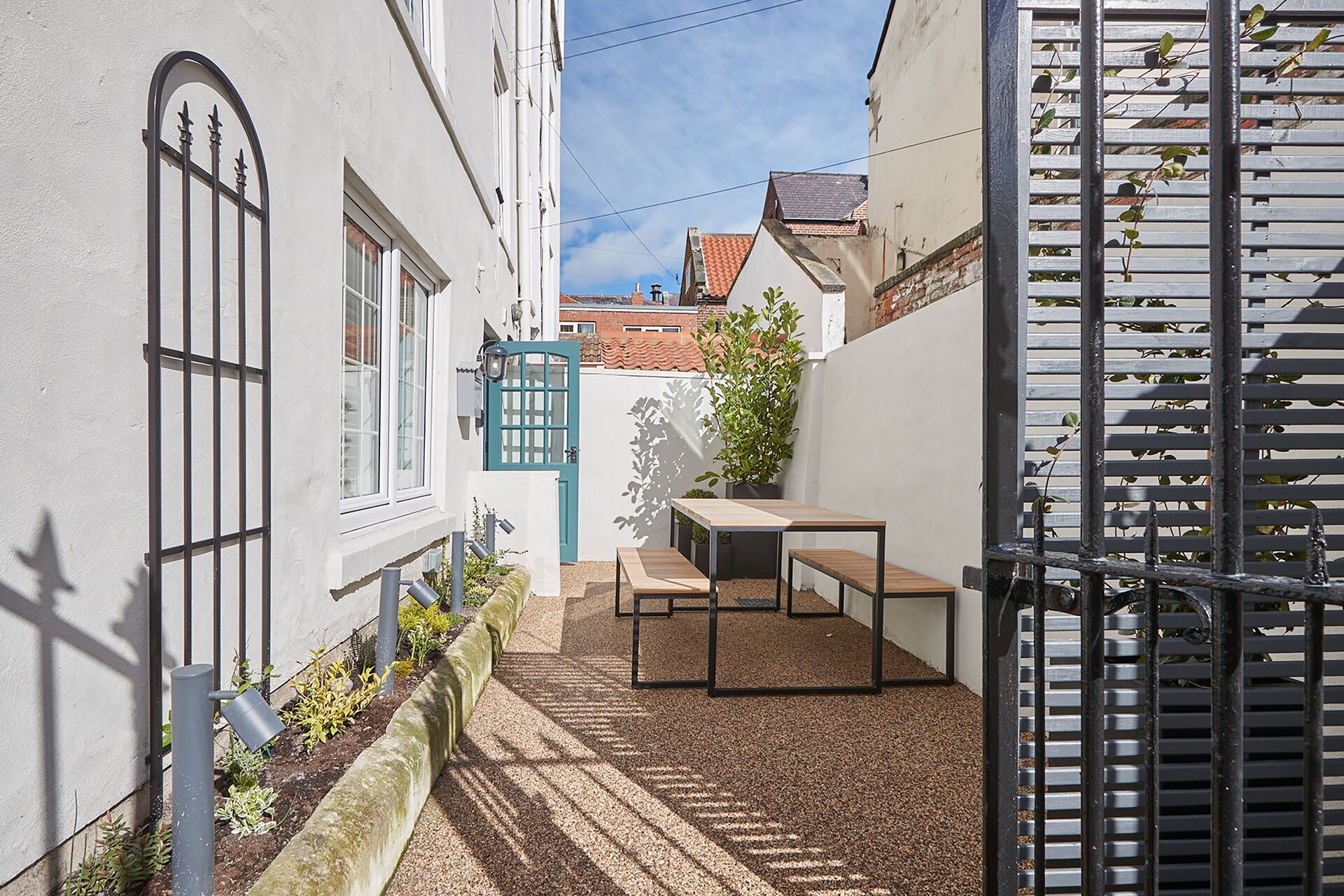 A narrow alleyway with a white building, a blue door, a small table, and a bench.