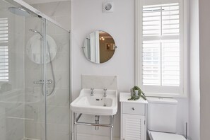 Un baño moderno con ducha envuelta en vidrio, un lavabo blanco con grifería cromada, un espejo redondo y una ventana con persianas.