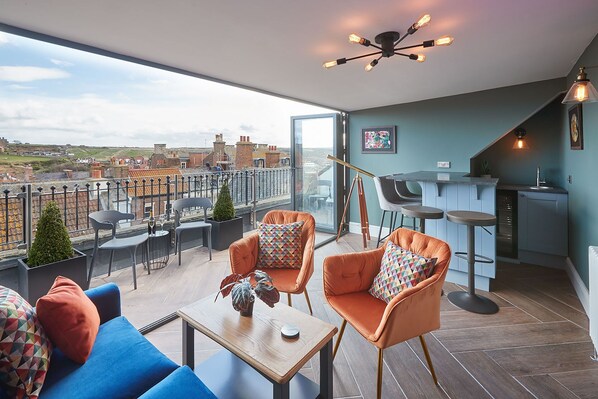 Un apartamento moderno con balcón, amueblado con sillas naranjas y un sofá azul, una pequeña zona de bar y un telescopio.