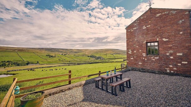 A rustic brick building with a small window, a wooden fence, and a gravel patio with a picnic table and benches. - Host Stay The Red Brick Barn (Bishop Auckland)