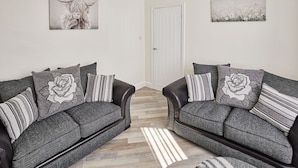 A living room with two grey sofas, each with rose-patterned pillows, a white door, and two wall-mounted artworks depicting a bull and flowers. - Host Stay 1 Montrose Street (Saltburn-by-the-Sea)