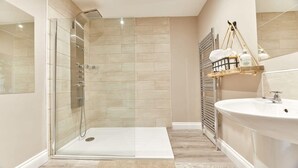 A modern bathroom with a glass-enclosed shower, a white sink, and a wooden shelf with toiletries. - Host Stay The Pepper Pot (Alnwick)