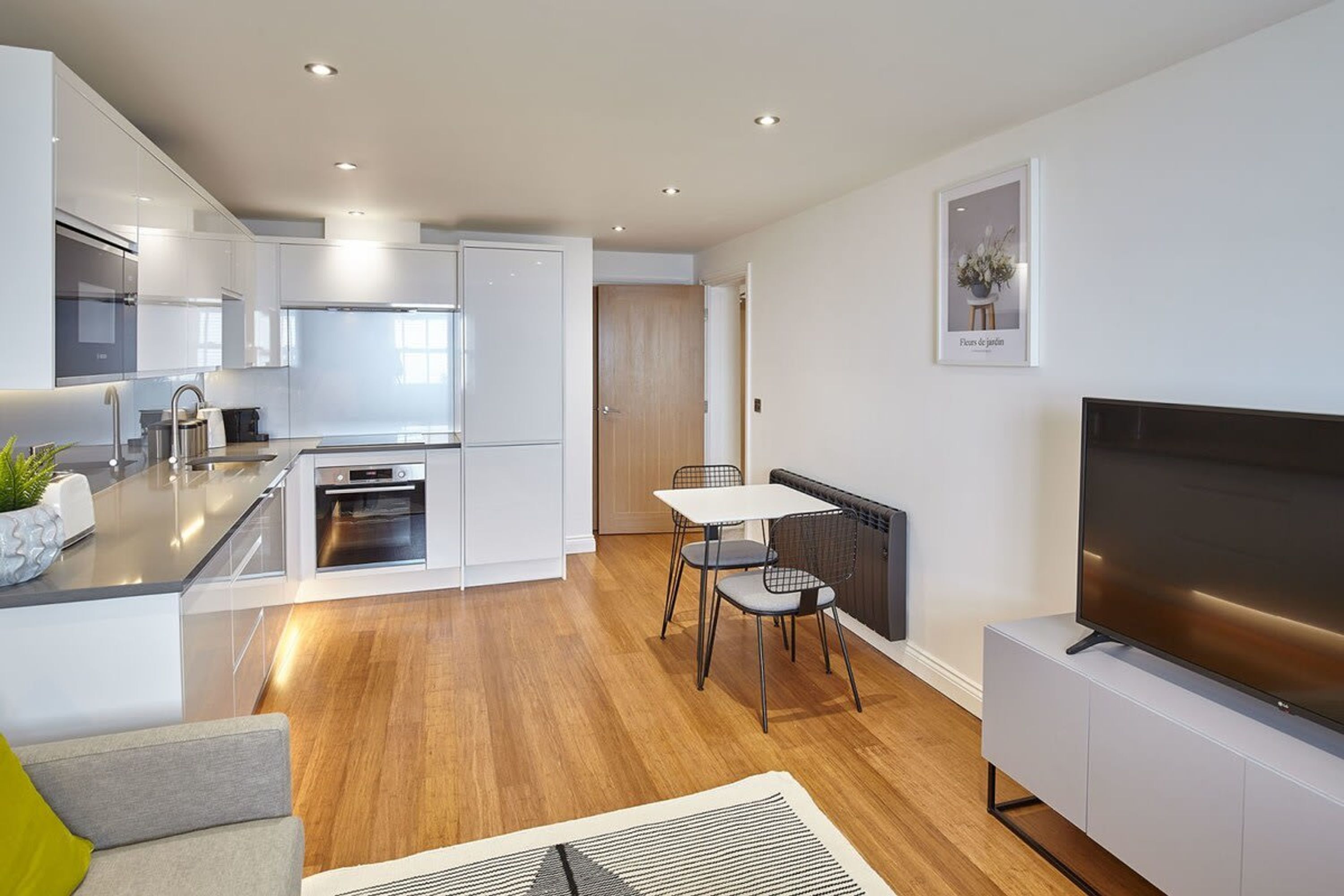 A modern kitchen with white cabinets, a dining area with a table and chairs, and a large flat-screen TV on a white stand.