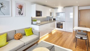 A modern kitchen with white cabinets, a built-in oven, and a microwave. There is a dining area with a white table and black chairs. - Host Stay Sandcastles Apartment (Whitby)