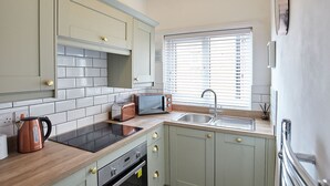 A compact kitchen with light green cabinets, a black induction cooktop, a built-in oven, and a window with blinds. - Host Stay Cobblestones (Redcar)
