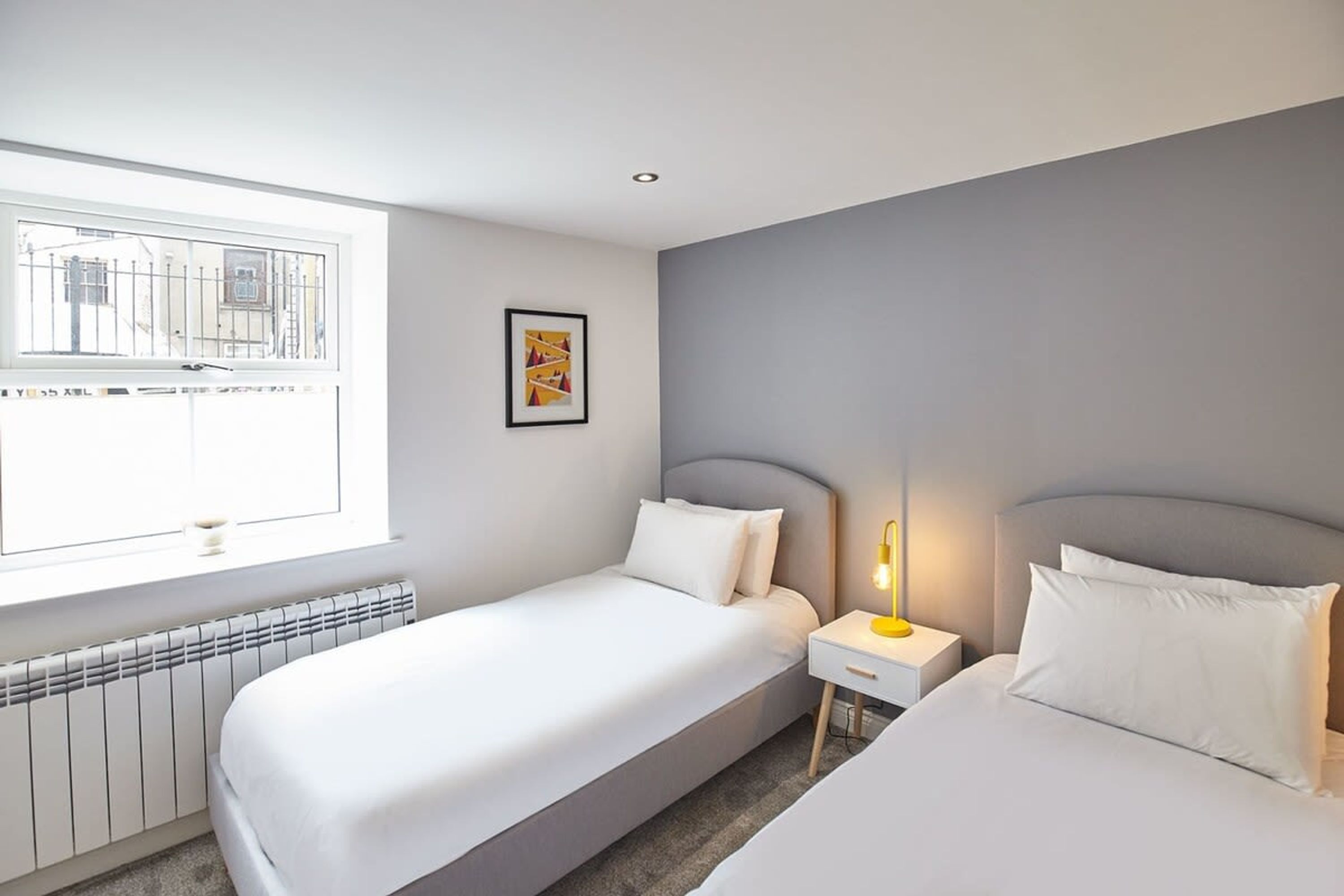 A hotel room with two beds, a window with a view of buildings, a radiator, and a bedside table with a lamp.