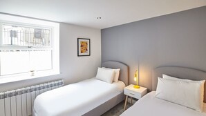 Quarto de hotel com duas camas, uma janela com vista para prédios, um radiador e uma mesa de cabeceira com abajur.
