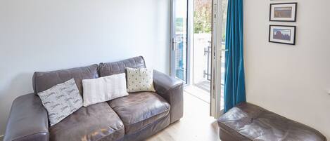 A living room with a brown leather sofa, a matching ottoman, and a glass door leading outside. There are three framed pictures on the wall.
