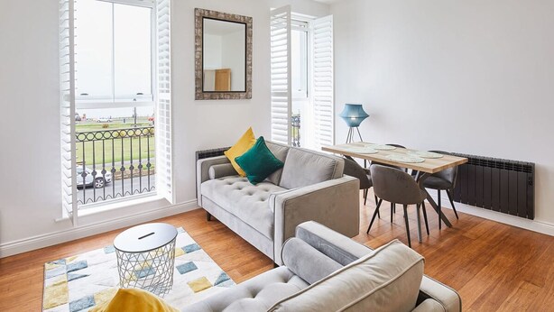 A modern living room with a grey sofa, a dining table with chairs, a small round table, and a view of the outside through large windows.