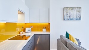 A modern kitchen with white cabinets, a yellow backsplash, and a grey sofa with a painting above it.