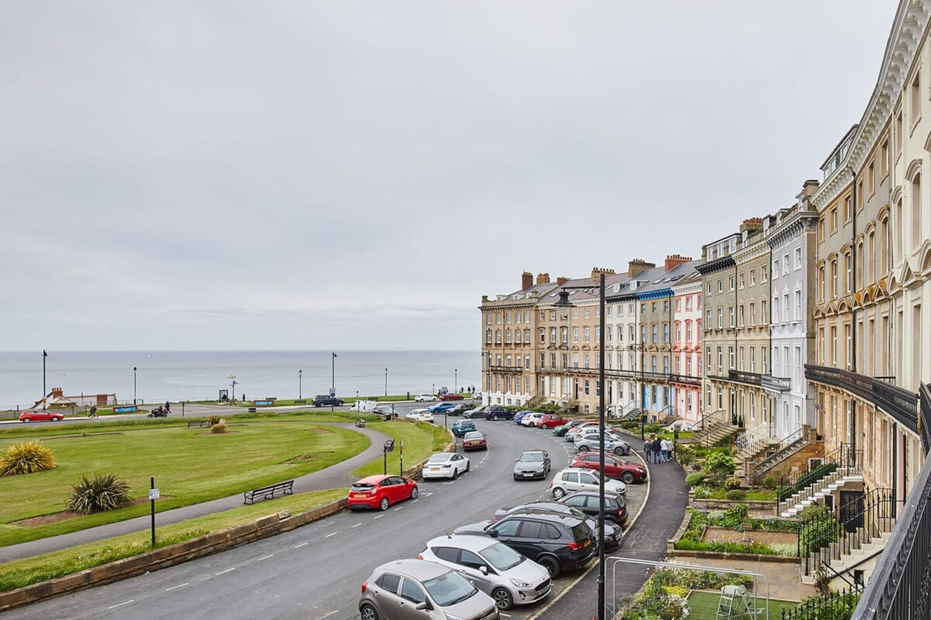 A coastal street lined with terraced houses, a parking area, and a grassy area with benches.