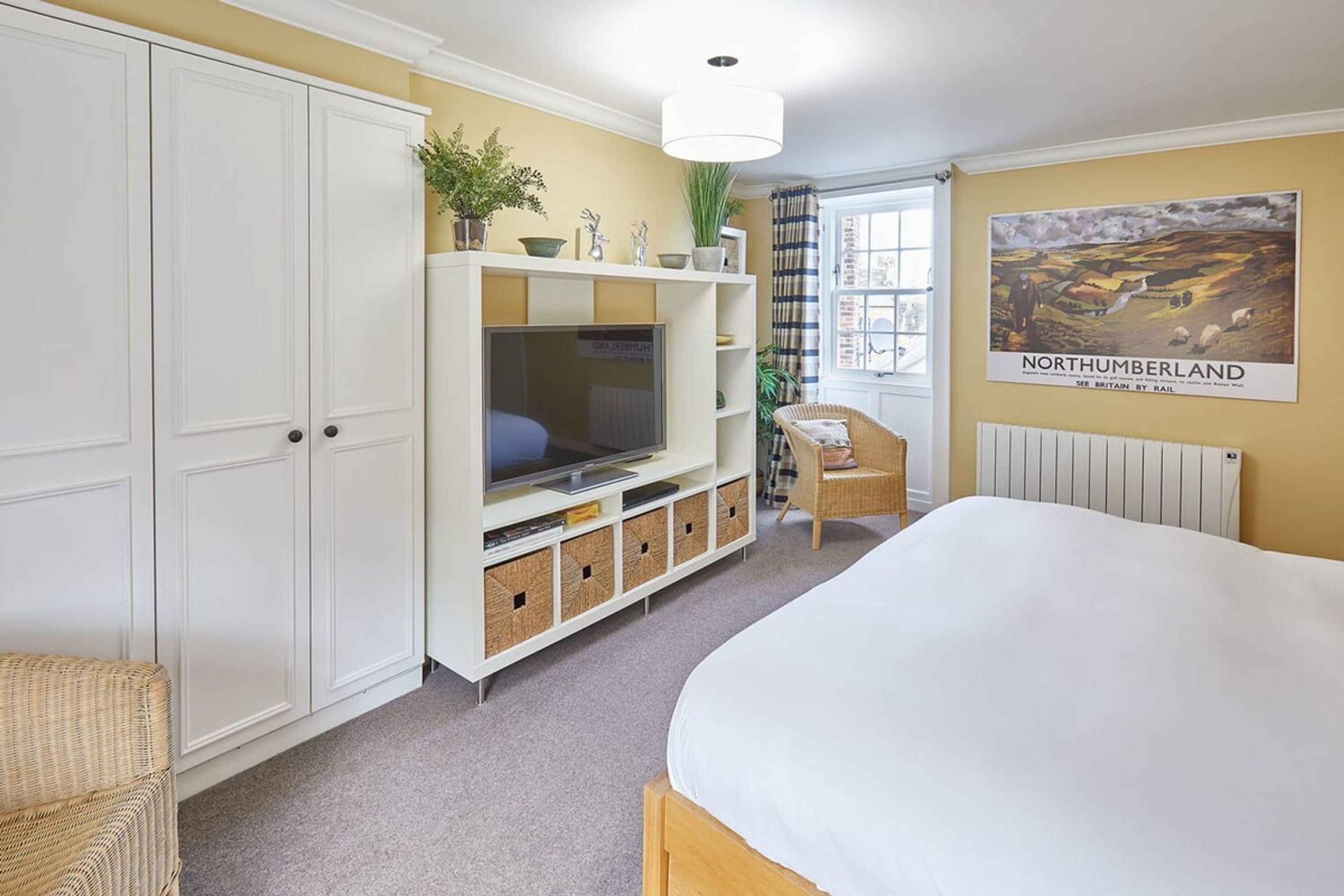 A bedroom with a bed, a TV on a white cabinet, a wicker chair, and a poster of Northumberland on the wall.