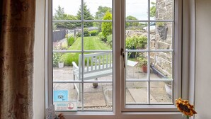 A view through a window with a white frame, revealing a garden with a white bench, potted plants, and a stone wall. - Host Stay Honeysuckle Cottage (Saltburn-by-the-Sea)