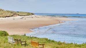 A beach with a wooden bench and a sign, a sandy shore, and a ship in the distance. - Host Stay The Puffins Nest (Seahouses)