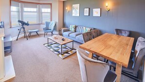 A cozy living room with a large window, a telescope, a sofa, a coffee table, and a dining table. - Host Stay The Puffins Nest (Seahouses)