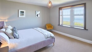 A bedroom with a large bed, a chair, a window with a view of the sea, and a unique geometric light fixture. - Host Stay The Puffins Nest (Seahouses)