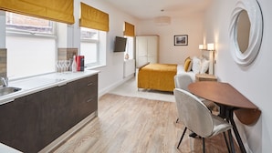 Quarto de hotel compacto com cozinha compacta, uma cama com colcha amarela mostarda, uma mesa de cabeceira com abajur e um espelho grande na parede.