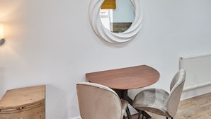 Uma área de refeições com uma mesa redonda, duas cadeiras, um baú de madeira e um espelho fixado na parede.