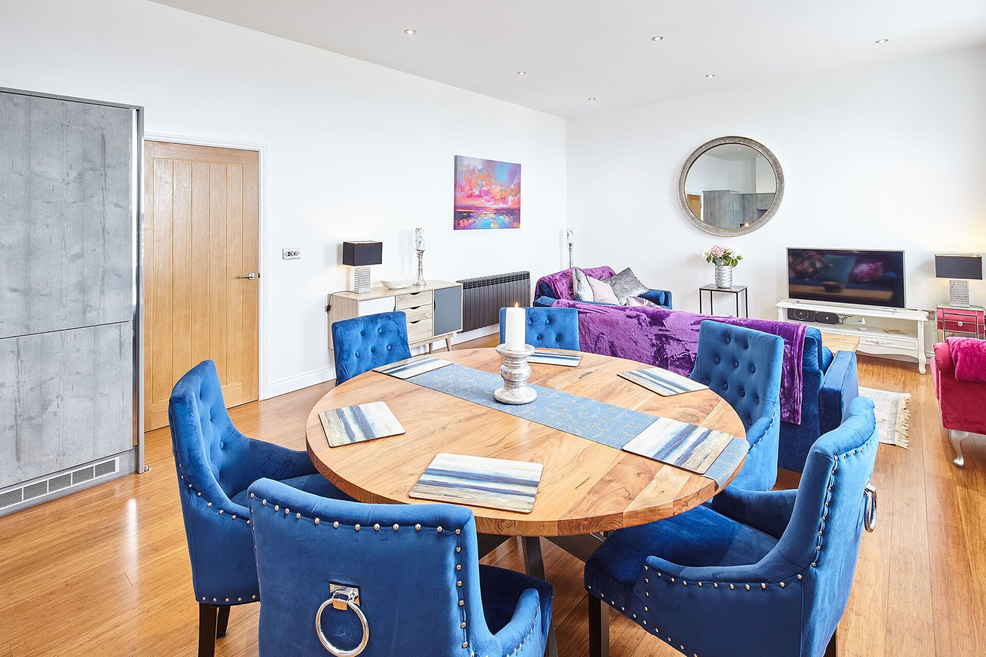 Un salon moderne avec une table de salle à manger ronde en bois, entourée de chaises à revêtement bleu agrémentées d’anneaux en métal. Un grand miroir est accroché au mur, et un téléviseur à écran plat est visible au fond.