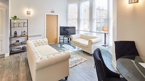 A modern living room with a beige sofa, a black armchair, and a glass coffee table. There is a TV on a stand, a bookshelf, and a window with blinds. - Host Stay Three Bays (Scarborough)