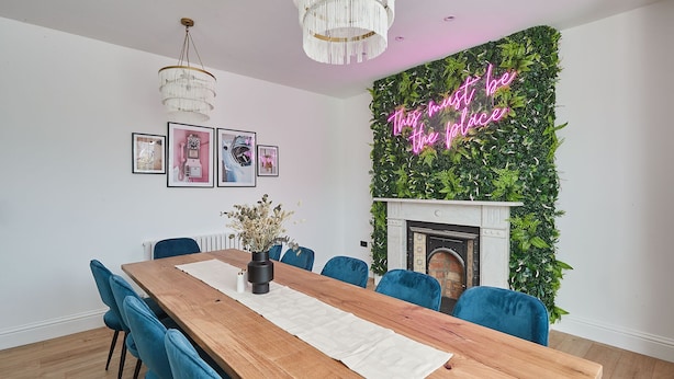 Une salle à manger avec une table en bois, des chaises bleues, un lustre, des tableaux encadrés et un panneau lumineux sur un mur vert.