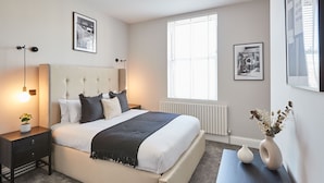 Une chambre moderne avec un grand lit, des tables de chevet, une petite table, une lampe sur pied et une fenêtre avec des stores.