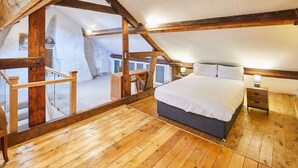 A bedroom with a sloped ceiling, wooden beams, a bed, bedside tables, and a view of a snowy outdoor area. - Host Stay The Duck Pond (Durham)
