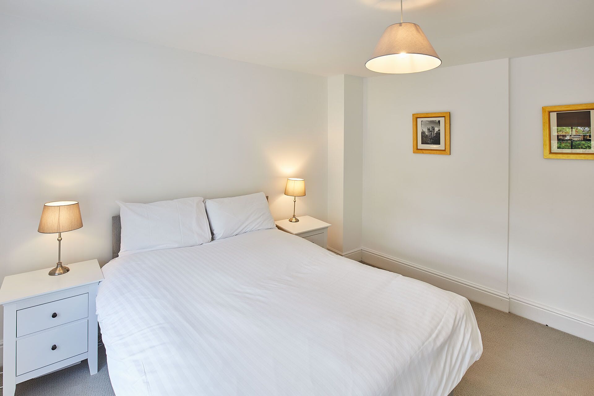 A neatly made bed with white linens, two bedside tables with lamps, and two framed pictures on the wall.