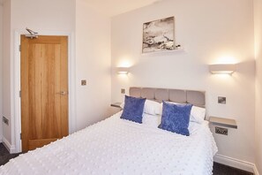 A bedroom with a wooden door, a bed with white bedding and blue pillows, two bedside tables with wall-mounted lamps, and a painting on the wall. - Host Stay Apartment 2 Queens Court (Scarborough)