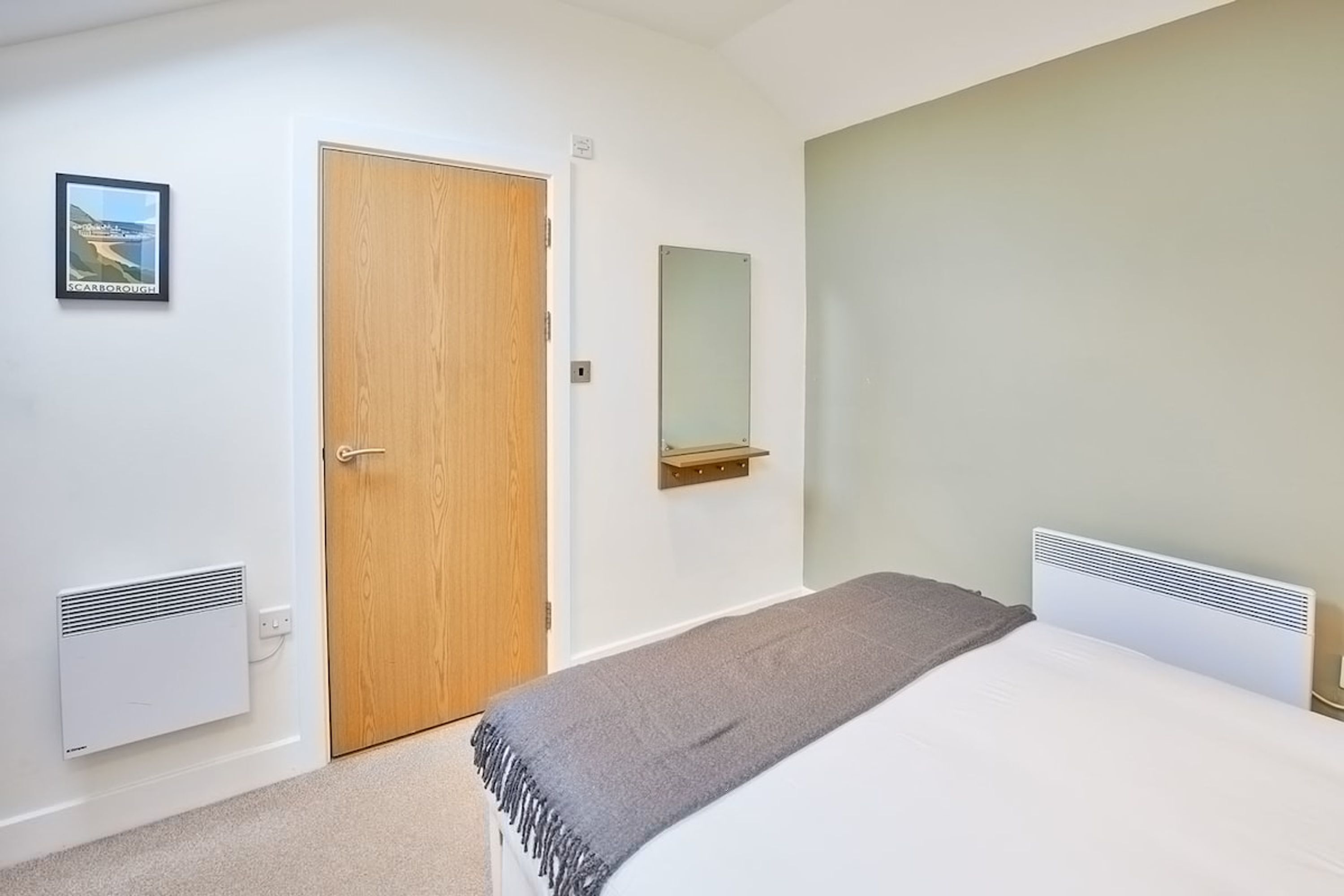 A bedroom with a wooden door, a wall-mounted mirror, a bed with a white headboard, and a radiator.
