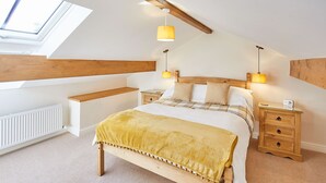 Quarto com teto inclinado, claraboia, cama com manta amarela, duas mesas de cabeceira com luminárias e cabeceira de madeira.