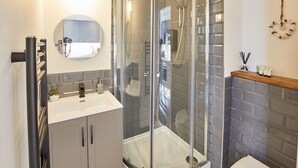 A modern bathroom with a glass-enclosed shower, a round mirror, and a wooden shelf with toiletries.