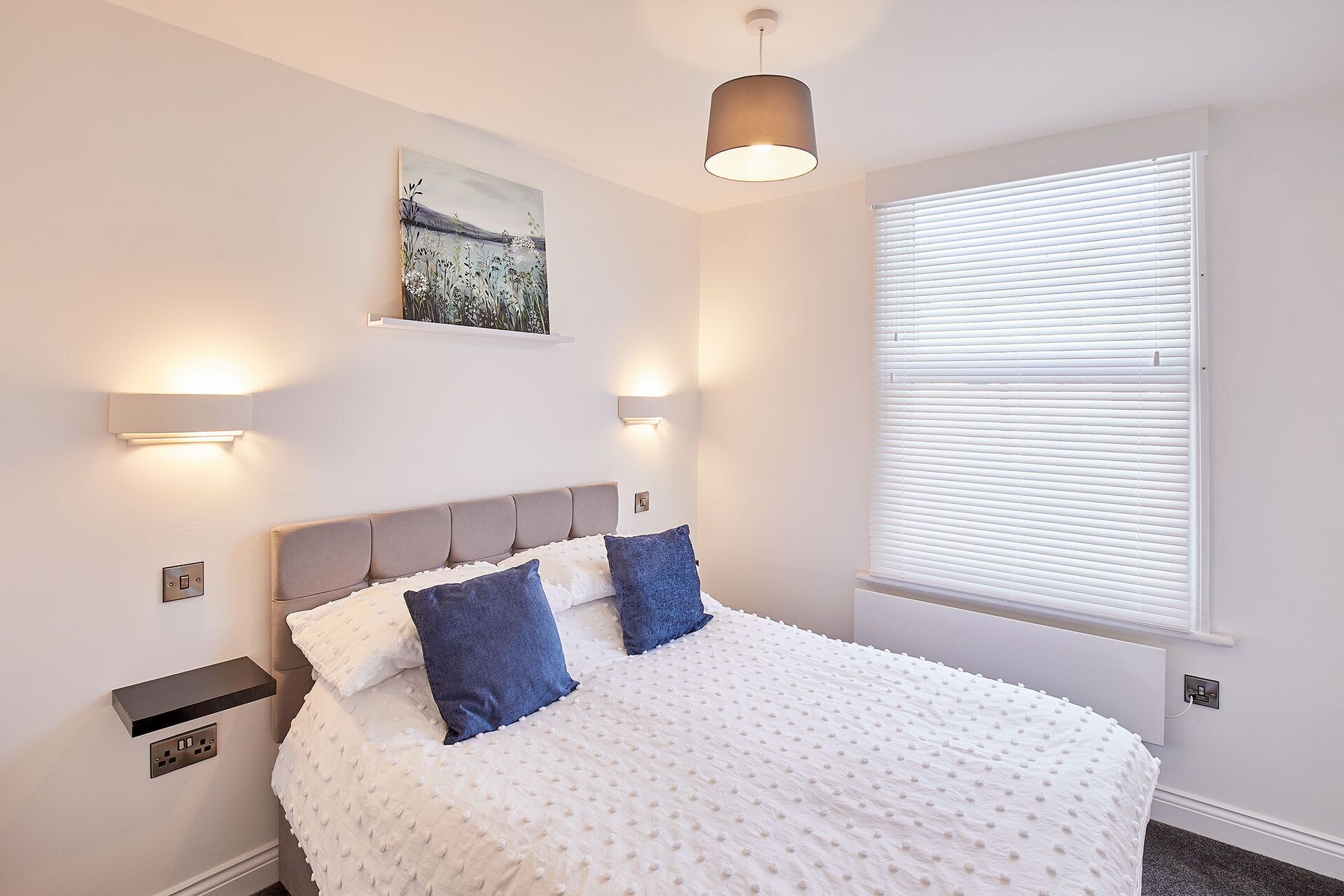 A bedroom with a bed, a painting, wall-mounted lights, and a window with blinds.