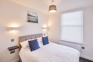 A bedroom with a bed, a painting, wall-mounted lights, and a window with blinds.