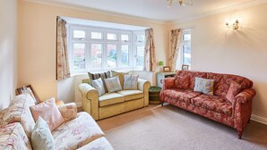 Ein gemütliches Wohnzimmer mit zwei Sofas, ein gemustertes rotes Sofa und ein beiges Sofa mit floralen Kissen. Es gibt Fenster mit Vorhängen, eine Chandelüster und einen kleinen Tisch mit einer Lampe.
