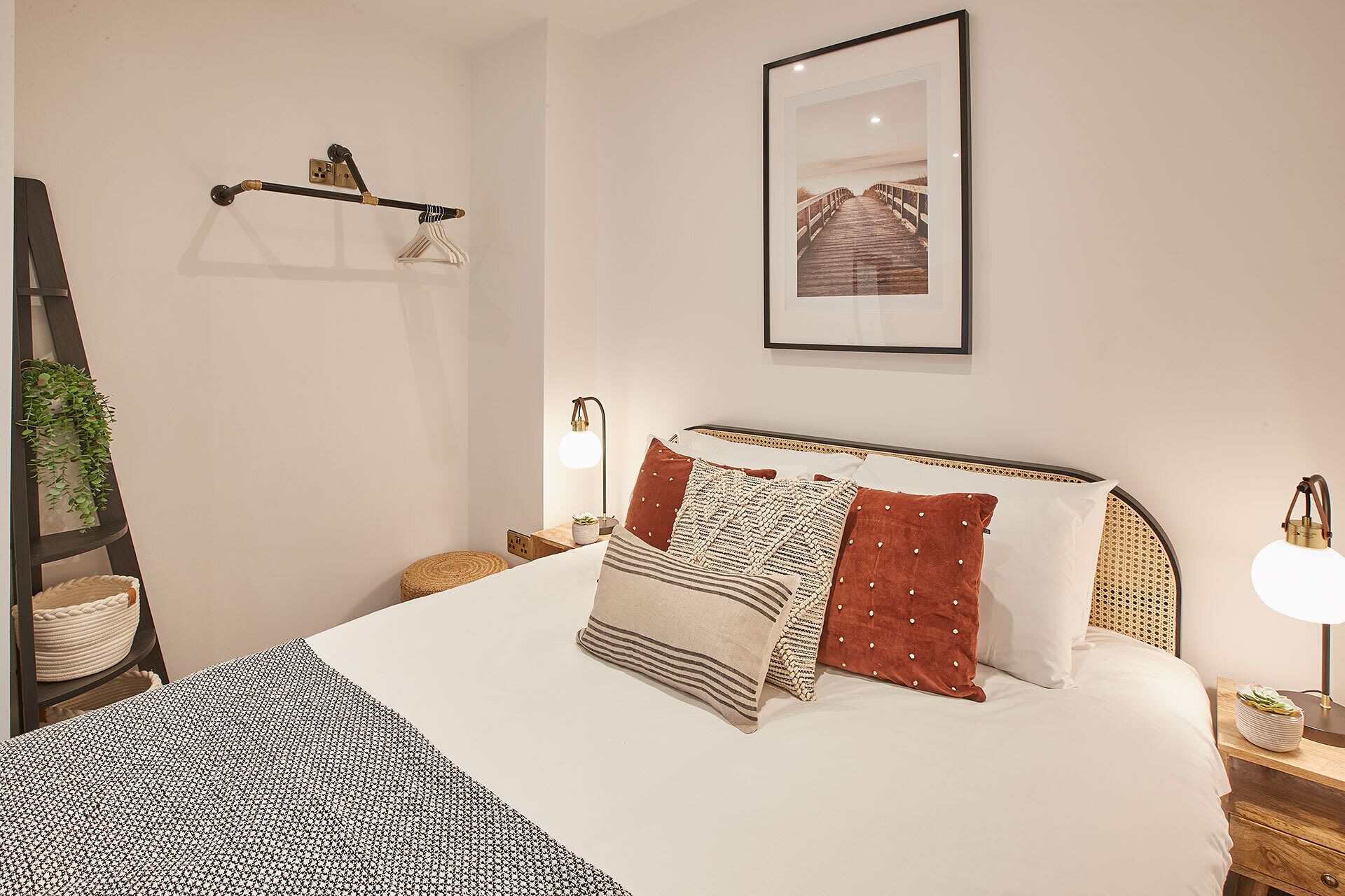 Une chambre bien rangée, avec un lit, des oreillers, une table de chevet, une lampe, un tableau encadré au mur et une plante suspendue.