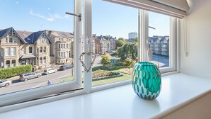 Een raam met uitzicht op een straat met traditionele huizen, een vaas op de vensterbank, en een helderblauwe lucht.