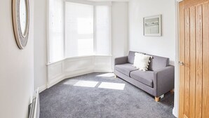 Une pièce avec un tapis gris, un canapé gris orné d’un coussin blanc, une porte en bois et une fenêtre avec des stores blancs.