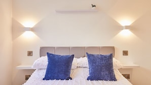 Un lit bien fait, avec une literie blanche et deux oreillers bleus, de part et d’autre de luminaires fixés au mur.