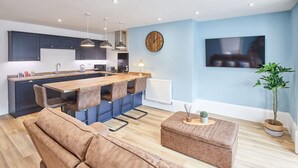 A modern kitchen with a central island, dark cabinets, pendant lighting, a clock, and a TV on the wall. - Host Stay The Seaside Escape (Saltburn-by-the-Sea)