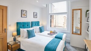 A double bed with teal headboard, white bedding, a bedside table with a lamp, a tray with drinks, and a window with blinds. - Host Stay The Seaside Escape (Saltburn-by-the-Sea)